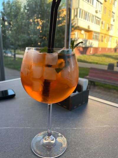 A close-up, eye-level shot shows a stemmed glass filled with an orange beverage, ice cubes, and mint leaves. Two black straws extend from the drink. The glass sits on a grey, speckled surface. In the background, through a window, a blurred green tree is visible, along with a bright yellow building with many windows and air conditioning units. A red and brown striped bench sits on a paved area outside. The scene appears to be an outdoor seating area in the city of Rozsoshentsi, Ukraine. A black mobile phone is also visible on the table to the left of the glass. A wooden holder with napkins is positioned behind the glass.