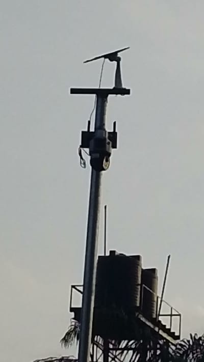 A tall metal pole stands against a pale sky, supporting a security camera under a solar panel. The pole has horizontal supports at its mid and upper sections. Behind it is a water tower with two black tanks on a metal framework, accessed by stairs. The image appears to have been taken in Enugwu-Ukwu, Nigeria. The ground is obscured by the lower portion of the water tower structure and some vegetation.
