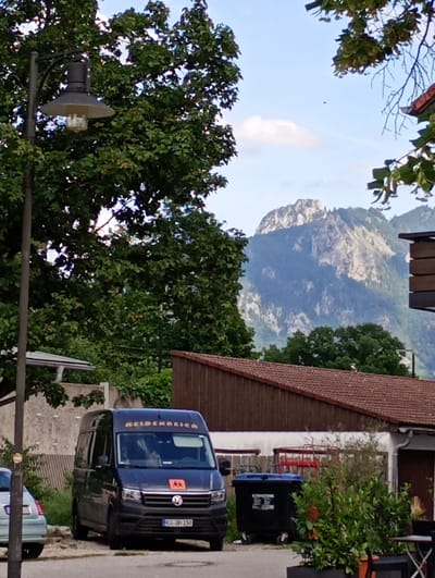 The image depicts a street scene in Brannenburg, Germany, with a black Volkswagen Crafter van parked on the right side. The van has the text "HEIDENREICH" in orange letters on its upper front, and a red circular logo with two figures. Its license plate is R-DH 193. To the left of the van, a light blue car is partially visible. In the background, a brown building with a red-tiled roof is situated behind a low wall and fence. A large black trash bin with a blue lid is also present. Further in the distance, a majestic mountain range rises against a partly cloudy sky. Lush green trees frame the scene, with one prominent tree on the left side of the image featuring a streetlamp attached to its trunk. The overall atmosphere is that of a quiet, somewhat rural or suburban setting within Brannenburg.