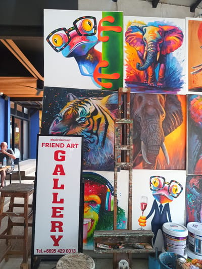 This outdoor scene in Ban Ao Nang, Thailand, displays a collection of colorful paintings exhibited outside a gallery named "Friend Art Gallery." The paintings predominantly feature wildlife like elephants and tigers, along with whimsical depictions of animals such as frogs with glasses and a monkey wearing headphones. A sign with the gallery name and contact information is prominently displayed. A man sits outside the gallery. A paint-splattered easel and supplies further suggest the artistic nature of the location.