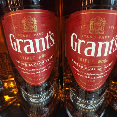 Two bottles of Grant's Triple Wood Blended Scotch Whisky are visible in the foreground. The labels are predominantly red with gold lettering and feature the Grant's crest. The bottles are dark amber glass and appear to be arranged in a retail setting. The background is dark and out of focus, with some glints of light. There are no people or animals present. The image does not provide any information about the location in the city of Los Teques, Venezuela.