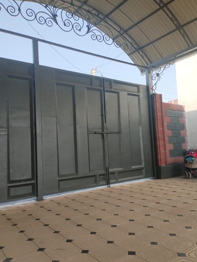The image shows a large, dark gray metal gate in the city of Октябрь, Kyrgyzstan. The gate has a structured design with rectangular panels and a horizontal locking mechanism. The gate is set within a paved area with interlocking tan tiles, each containing a small dark square in the center. To the right of the gate is a short pillar constructed of red and dark gray bricks. A light-colored wall is visible to the far right. Overhanging the scene is a metal awning supported by decorative scrollwork. In the background, the sky is clear and blue, with some utility poles and wires visible. A baby stroller is parked to the right of the brick pillar, with some items inside. The overall impression is that of an entrance to a residential or commercial property.