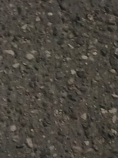 The image shows a close-up, abstract view of a dark, granular surface, possibly asphalt or concrete with aggregate. The texture is rough, with visible small stones or pebbles embedded in a matrix. The lighting appears even and diffused, creating a lack of distinct shadows. There are no discernible features that would indicate a specific location within the city of Savino, Russia, or any people, objects, or actions. The image focuses solely on the material's texture and color.