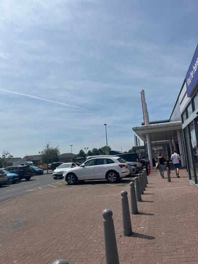 The image depicts a street scene in East Suffolk, United Kingdom. A line of parked cars is visible along a building, with a white car being the most prominent. Pedestrians walk along the paved area adjacent to the building. Bollards line the pavement. The sky is blue with some clouds. A sign reading "You're Here" is partially visible on the building.