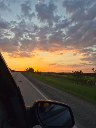 The image shows a view from the passenger side of a car traveling on a paved road during sunset. The sky is filled with a dramatic display of orange and yellow hues, with scattered dark clouds. The road curves to the left, with a grassy shoulder and trees lining the right side. In the distance, rolling hills are visible against the bright sky. The side-view mirror of the car is visible in the foreground, reflecting a portion of the interior and the passenger. The overall impression is one of a peaceful, scenic drive through the countryside of Забегалово, Russia.