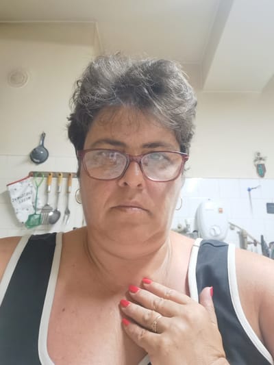 A middle-aged woman with dark, curly hair and glasses is shown in a close-up selfie. She is wearing a dark tank top with white trim. Her right hand, with bright red nail polish, is resting on her chest, and she is wearing a ring on her left hand. The background appears to be a kitchen with white tiled walls, cooking utensils, and a white water heater. The image does not provide any information about the city of Lomets, Bulgaria.