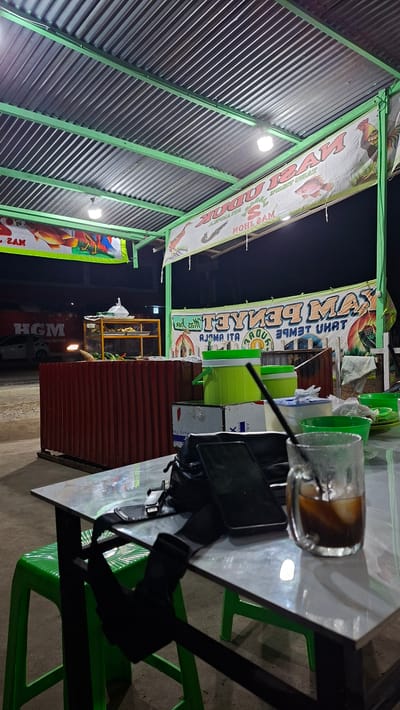 The image depicts an outdoor food stall at night in Keudebayu, Indonesia. The stall is covered by a corrugated metal roof supported by green metal poles. Banners displaying images of fish and chickens are hung from the roof. A glass of iced coffee sits on a table in the foreground, along with a black bag and a smartphone. Behind the table, there is a food display counter made of red corrugated metal, with a yellow metal shelf displaying various food items. Green plastic chairs are visible, and in the background, a white car is parked. The scene is lit by overhead lights, casting shadows and highlighting the details of the stall.