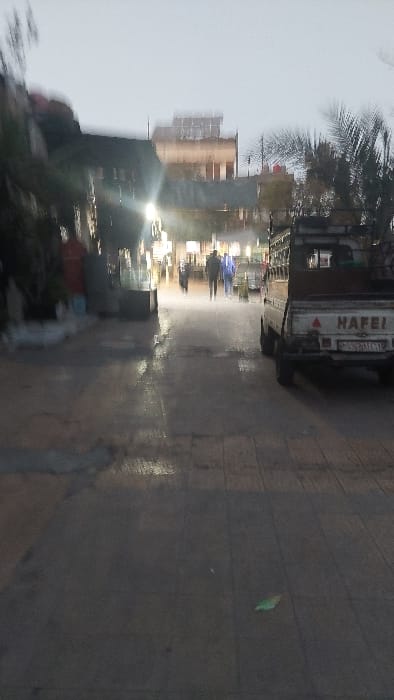 The image depicts an urban street scene within Al-Qaboun Municipality, Syria, under low light conditions, possibly after rainfall, evidenced by wet and reflective ground surfaces. The perspective is directed down a paved thoroughfare, flanked by buildings and commercial establishments. On the left, obscured elements consistent with storefronts or residential facades exhibit artificial illumination. On the right, a light-colored Hafei pickup truck with a utility bed is stationary or parked, its rear facing the viewer. The truck's tailgate displays the word "HAFEI" and a Syrian license plate with Arabic script and numerals. In the mid-ground, multiple human figures are observed walking away from the camera, proceeding towards a brighter, more densely lit section of the street. Distant background elements include additional buildings with visible windows or light sources, and several palm trees. The ground in the foreground exhibits paving with distinct tile lines, and a small, bright green object, possibly a leaf or debris, is visible on its surface. The overall image quality is blurred, consistent with motion or low-light photography.