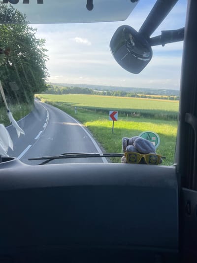 A gray plush bunny wearing yellow sunglasses with the Lidl logo sits on the dashboard of a vehicle, looking out at a rural road. The road curves to the left, bordered by green fields and trees. A road sign with a red chevron indicates a sharp left turn. In the background, a hazy landscape of rolling hills and farmland is visible under a bright, cloudy sky. The interior of the vehicle, including the windshield wipers and part of the dashboard, is visible in the foreground. No specific landmarks or buildings identifiable as being in Essen, Germany are present in this image.