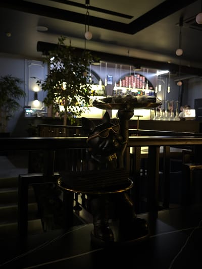 A whimsical scene unfolds in a restaurant setting. A dark, sculptural figure of a bull, donned with sunglasses, is centrally positioned. This figure sits atop a staircase or elevated platform, adding to its prominence. In the backdrop, a wine display is partially visible. A lush green bush provides a touch of nature to the interior. Soft lighting creates ambiance, highlighting the unique decor.