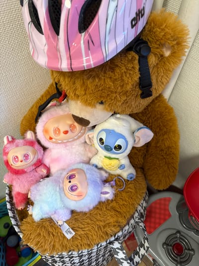 A medium shot shows a brown stuffed teddy bear wearing a pink and white striped helmet. The bear is holding several smaller stuffed toys, including a white Stitch character from Disney, and three fuzzy dolls with large eyes and distinct facial features in pink, purple, and pastel blue. The toys are nestled within the bear's arms and on top of a fuzzy brown surface, which appears to be part of the bear itself. The bear and toys are placed inside a woven basket with a black and white geometric pattern. The background consists of a textured wall. A red toy stove is partially visible to the right of the basket. The scene is indoors, with no specific landmarks in Oslo, Norway, identifiable.