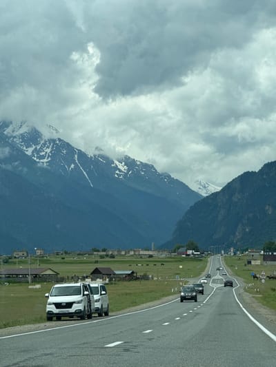 The image captures a scenic view of a road leading into a mountainous region near the city of Verkhnyaya Teberda, Russia. The foreground features a paved road with white lane markings, and on the shoulder, two white passenger vans are parked with their doors open. Several other vehicles, including cars and SUVs, are traveling along the road in both directions. The middle ground displays a vast green field dotted with scattered buildings, some appearing to be farm structures and houses. A herd of animals, possibly horses, is visible grazing in the field. In the background, imposing, dark blue mountains rise dramatically, their upper reaches capped with snow and shrouded in cloudy mist. The sky is overcast with heavy, grey clouds, suggesting potentially cool or inclement weather. The overall impression is one of a rural, mountainous landscape with a road serving as a connection to a remote settlement.