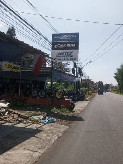 The image presents an outdoor commercial scene under clear, bright daylight in Beteng_Pangkhar_Somthrang, Bhutan. In the left foreground, a cracked concrete sidewalk is strewn with wooden debris and a crumpled blue plastic or fabric item. This leads to a single-story building, an apparent tire and wheel shop, identified by a prominent vertical sign displaying "ACCELERA," "FORCEUM," "ZEETEX," and "SYIFA JAYA BAN." The shop's open facade showcases numerous vehicle tires and alloy wheels, arranged on display stands and a low, red concrete structure. A red and white bicolour flag hangs vertically from the building's overhang, adjacent to a yellow sign indicating "Rp 25.000*." Numerous overhead utility lines crisscross the expanse of the light blue sky. To the right, an asphalt road extends into the background, where a dark sport utility vehicle and a smaller unidentified vehicle are observed traveling away from the camera. Sparse green foliage and trees line the far side of the road, with additional utility poles and wiring visible. No individuals are clearly discernible in the scene. Strong shadows are cast, indicative of direct sunlight.