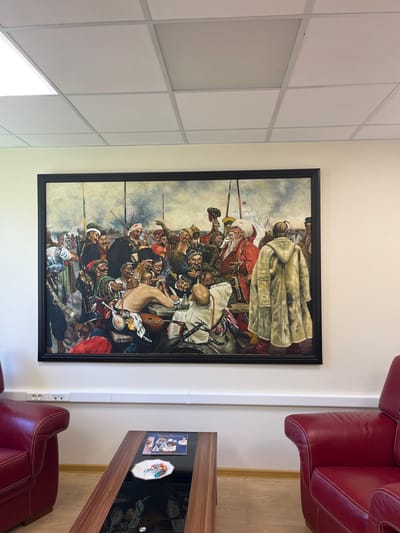 The image depicts a large, framed painting hanging on a plain, cream-colored wall in what appears to be an office or waiting area. The painting itself is a detailed, somewhat chaotic scene with numerous figures, likely historical or mythological, dressed in varied, traditional attire, some with weapons. They are gathered around a table, engaged in conversation or activity, with a backdrop of a cloudy sky and distant figures. Below the painting, there is a white electrical conduit running along the wall with several outlets. In the foreground, two plush red leather armchairs are visible on either side of a dark wood and glass coffee table. The coffee table has a magazine and a small decorative plate with wrapped candies on it. The flooring is light-colored wood. The ceiling features a grid of white acoustic tiles with recessed lighting. The overall impression is one of an interior space decorated with a significant piece of art, likely in a professional or public setting. No specific location in Geneva, Switzerland, can be determined from the image itself.