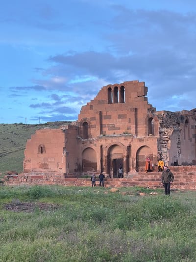 A horizontal, outdoor shot shows the ruins of a red stone structure with people standing in front of it. The structure is located in the city of Anipemza, Armenia. The ruined structure features several arched openings and a row of vertical pillars at its top. To the right of the structure is an expanse of green grass, and to the left is a blue sky with clouds. Several people are visible near the structure: one person is wearing a yellow jacket and another is wearing a red/orange outfit. A person wearing a dark jacket and pants is standing towards the right.