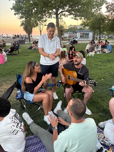 In Kadıköy, Turkey, a group of friends enjoys a pleasant evening outdoors on a grassy park. The setting sun casts a warm, orange glow across the sky, indicating it's late afternoon or early evening. The overall mood appears relaxed and joyful as people gather, some sitting on blankets and others in portable chairs.
The central focus is on a man playing an acoustic guitar, entertaining those around him. Nearby, a woman is clapping along to the music, a smile on her face, suggesting she's enjoying the performance. Other individuals are seen sitting, talking, and socializing, creating a convivial atmosphere. A variety of objects are visible, including the guitar, a phone, beverage cans, and picnic items like popcorn. The people are dressed in casual summer attire, with t-shirts, shorts, and sneakers being common.
The scene includes a glimpse of a building in the background, possibly a small kiosk or information booth, and trees providing shade. In the distance, more people are spread out across the park, some walking or sitting by what appears to be a waterfront. The presence of a dog adds to the relaxed, community feel of the gathering. The weather seems to be clear and warm, suitable for an outdoor social event. There is some visible text on the t-shirt of the person in the foreground, but it is not fully legible.