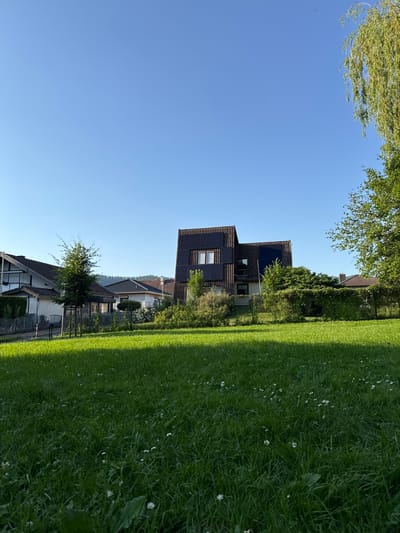 This is a landscape image captured outdoors on a bright, sunny day, likely in the afternoon, given the strong shadows. The scene depicts a residential area with a focus on a modern, multi-story house featuring a dark facade and solar panels on its roof and walls. The house is situated amidst lush green grass and trees, suggesting a well-maintained suburban or rural environment.

In the foreground, a vibrant green lawn stretches across the left side of the frame, dotted with small white wildflowers. The grass is neatly mowed, and a distinct shadow line indicates the presence of a bright light source, consistent with a sunny day. Behind the lawn, a fence encloses part of the property.

To the right of the lawn, the modern house stands out with its dark, possibly wooden or composite cladding. The solar panels are prominently visible, indicating an eco-conscious design. The house has several windows, some of which are reflecting the blue sky. Adjacent to this house, other residential buildings with traditional tiled roofs are visible, suggesting a mix of architectural styles in the neighborhood.

The background is dominated by a clear, bright blue sky, with no clouds visible. Trees with green foliage are scattered throughout the scene, contributing to the natural setting. In the far distance, a tree with light-colored, possibly wind-swept branches is visible against the sky.

There is no visible text within the image. The overall mood of the image is peaceful and serene, highlighting a well-kept property on a beautiful day in Bevern, Germany.
