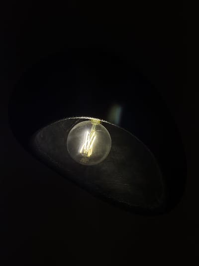 An Edison-style incandescent light bulb glows brightly within a dark, conical lampshade. The bulb's filament, a series of thin, vertical wires, is clearly visible and emits a warm yellow light. The lampshade is a deep black with a textured, matte finish that absorbs most of the light, creating a stark contrast with the illuminated bulb. The background is entirely black, suggesting a dimly lit or nocturnal setting. The image is focused on the light bulb and the immediate vicinity of the lampshade, with no other objects or people present. The lighting suggests a single, localized light source. The image is a close-up shot, emphasizing the details of the light bulb and the texture of the lampshade. The scene is devoid of any environmental cues that would indicate a specific location within Sithonia Municipal Unit, Greece, or any other geographical area.
