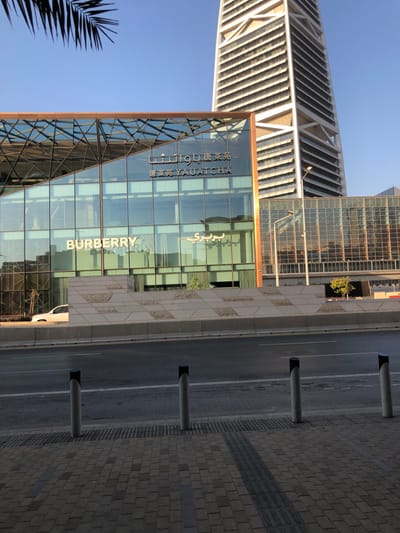 A tall, modern building with glass windows showcasing "BURBERRY" and other text in multiple languages. Adjacent to the building, a distinctive, partially visible architectural structure with a series of horizontal lines. A paved walkway runs along the side of the building, separated from the road by a row of bollards. A palm tree is visible in the lower left corner. The composition is shot from a low angle, emphasizing the height of the structures.