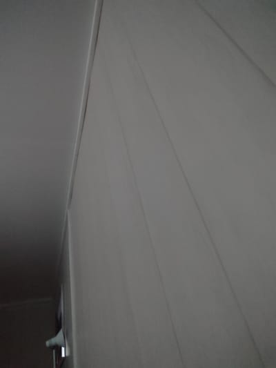 The image depicts an interior view of a room with plain, light-colored walls. The left side of the image shows a darker area, possibly a ceiling or a wall, with a distinct straight seam or edge where it meets the lighter wall. This seam runs diagonally from the top left towards the bottom right. The dominant feature is the textured, possibly paneled, wall on the right, which has subtle vertical lines or creases that create a subtle pattern of light and shadow, suggesting a draped or folded surface. In the bottom left corner, a small portion of an object is visible, appearing to be white and protruding from the darker area. This object is partially obscured and its nature is unclear, but it might be a light fixture or part of some equipment. The overall lighting is dim and diffused, with no direct light sources visible. The scene is static and shows no activity or people. The location within Қосшағыл ауылдық округі, Kazakhstan is not visually indicated in the image.