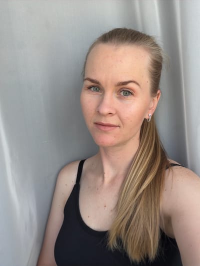 A close-up, head-and-shoulders shot of a fair-skinned woman with blonde hair pulled into a ponytail, wearing a black tank top, and silver hoop earrings. She has light green eyes and freckles scattered across her cheeks and shoulders. The background consists of a neutral, grey-toned fabric that appears to be a curtain or backdrop. The lighting is soft and diffused, creating a natural look. The woman is positioned slightly to the left of the frame, looking directly at the camera with a neutral expression. The environment suggests an indoor setting, likely a studio or a well-lit room. There are no visible contextual clues to indicate the specific location within the city of Кульдур, Russia.