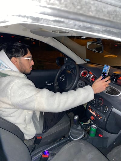 An individual with dark hair and a beard is inside a car. The person wears glasses and a white jacket. They are wearing a seatbelt and have a mobile phone mounted on the dashboard. The interior of the vehicle is visible, showing the steering wheel, dashboard controls, gear stick, and a beverage can. Outside the car, a dark background suggests nighttime or an indoor setting.