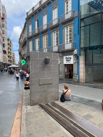 The image captures a street scene in Las Palmas de Gran Canaria, Spain. A four-story blue building with white-framed windows and decorative wrought iron balconies stands to the left of the frame, merging with a modern glass structure above. To the right of the building's ground level is a "Flying Tiger Copenhagen" store.
A large stone monument with a bronze head affixed to it is centrally positioned, acting as a focal point. People are visible on the sidewalk, including a woman sitting on the edge of a raised platform near the monument, appearing to use her phone. Pedestrians are walking further down the street, and a green pharmacy sign is visible in the distance. The sky is partly cloudy, and other buildings line the street.