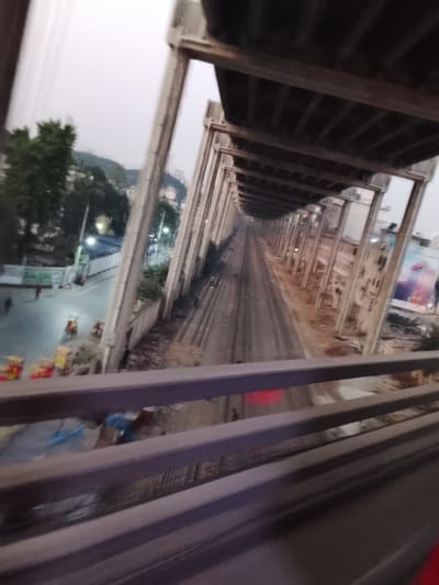 A train track stretches into the distance, enclosed by a concrete structure with regular vertical supports and a covered top. The perspective is from a high angle, likely a bridge or overpass. To the left, a street scene with buildings, trees, and people is visible. On the right, a building with advertising posters is apparent. The overall atmosphere is somewhat hazy and gray. The foreground features the railing of the structure from which the photograph is taken.
