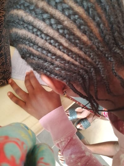 A close, high-angle shot shows a person with braided hair. The braids are black and brown and neatly organized. The person's right hand is visible, with tanned skin and slender fingers resting on a piece of paper with writing on it. The person is wearing a light pink, long-sleeved top. The bottom of the frame shows blue and gray sandals and a part of the person's legs. A small table is present, with an object wrapped in patterned fabric partially visible in the bottom left corner. The scene appears to be indoors, possibly in Ogboko, Nigeria.
