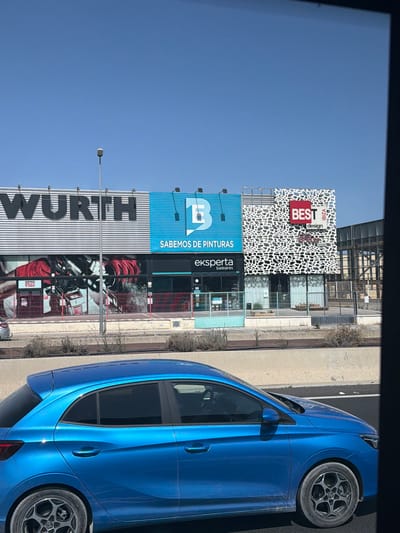 The image shows a bright blue car driving on a road with a commercial building in the background. The building has several storefronts with prominent signage. One sign reads "WURTH" in large black letters. Another section of the building has a blue sign with a white "B" logo and text that appears to say "SABEMOS DE PINTURAS". Next to this is a storefront with a distinctive white decorative facade and a red sign that says "BEST" with "design" underneath, and "IBIZA" above. Below "BEST" is a smaller logo that appears to be Adidas. The storefronts are behind a light-colored fence. The sky is clear and blue. The overall scene suggests a commercial area, likely in Sant Antoni de Portmany, Spain, given the "IBIZA" signage.