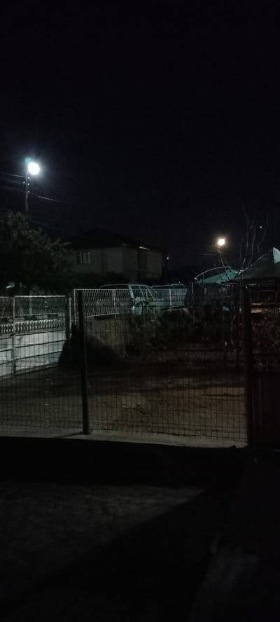 This is a nighttime photograph of an outdoor scene, likely in Iskra, Bulgaria. A tall street lamp illuminates a portion of the foreground and middle ground. Beyond the lamp post, a residential building with visible windows is partially visible in the background. A fence, composed of metal mesh and vertical posts, runs across the middle ground. Behind the fence, several vehicles, appearing to be vans or minivans, are parked. There are also some dark, dense bushes and possibly some parked equipment or structures behind the vehicles. The overall environment is dark, with the street lamp providing the primary source of light. The ground in the foreground is dark and appears to be paved or covered in dirt, with shadows cast from the fence.