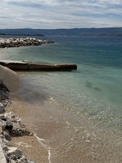 The image shows a view of the Adriatic Sea from the beach in Dugi Rat, Croatia. The scene is captured from the shoreline, looking out towards the water. The foreground features a sandy beach with a concrete platform extending into the shallows. Several large rocks are visible in the foreground, and the water is clear, with light ripples on the surface. In the background, the coastline stretches out towards the horizon, and the water becomes darker and more opaque, with a slight chop. The sky is partly cloudy, with bright blue patches and wispy white clouds. There are no people visible in the image.