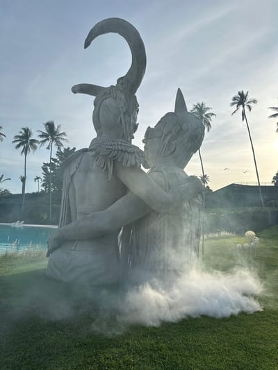 This is a vertical, eye-level, medium shot of two large concrete sculptures in an outdoor setting with a pool and grassy area. The sculptures appear to depict two figures embracing, possibly in a mythological or artistic representation. One sculpture has large horns curling downwards and backwards, while the other has a more human-like profile with feathered adornments around its neck and shoulders. The figures are rendered in a detailed, stylized manner, suggesting a contemporary art installation.
The scene is illuminated by what appears to be daylight, with the sun low in the sky, casting a soft glow. There is a noticeable haze or fog around the sculptures, particularly on the right side of the frame, obscuring some details and adding a dramatic effect. In the background, there are several tall palm trees, characteristic of a tropical location. Beyond the trees, there is a modern building structure. The ground is covered in lush green grass and a bright blue swimming pool is visible in the lower portion of the image.
The overall mood is somewhat mysterious and ethereal due to the fog and the abstract nature of the sculptures. The embracing figures and the tropical setting evoke a sense of serenity or perhaps a dramatic narrative. There is no visible text in the image. The location context of Baan Lipa Noi, Thailand, reinforces the presence of palm trees and the likely warm, humid climate.