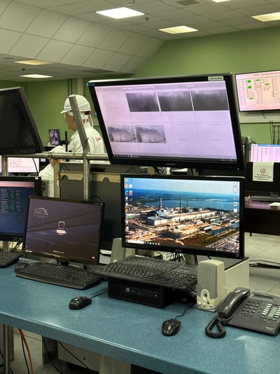 This image depicts a brightly lit control room located within the Chernobyl Nuclear Power Plant, near the city of Pryp'yat', Ukraine. The room features light green walls and a white grid ceiling with recessed square fluorescent lights.

In the left background, a male individual is partially visible, facing right. He wears a white hard hat with a red device on the back and a white uniform shirt or lab coat with a partially obscured red logo on the right chest area. He appears to be seated or standing at an adjacent workstation.

The foreground is dominated by a blue-green speckled countertop holding multiple computer workstations. From left to right: a black keyboard and mouse are positioned on the left. In the center, a compact black HP desktop computer tower (HP EliteDesk 800 G1 Desktop Mini) sits beneath a black keyboard and mouse. To the right of the HP tower is a beige and gray speaker system with a green power indicator light. Further right is a black landline telephone with a digital display and numeric keypad.

Several monitors are visible. On the desk level, three monitors are arranged: one partially visible on the far left displaying technical data, a central monitor with a black bezel displaying a dark screen with a white stylized computer icon and the text "SMEBEC", and an LG monitor on the right displaying an aerial photograph of the Chernobyl Nuclear Power Plant as its desktop background, showing multiple reactor buildings, cooling towers, and a large body of water.

Above these, a large Samsung monitor is mounted, displaying a grid of six frames. Four frames show dark, grainy imagery, possibly from cameras or sensors, while two frames are blank. To the far right in the background, another monitor displays a control panel schematic with numerous green and red indicators and Cyrillic text, including "ОБКВ - Система Вентиляции" (OBKV - Ventilation System). A white sign with a red circular 'no' symbol and Cyrillic text is also partially visible in the right background.