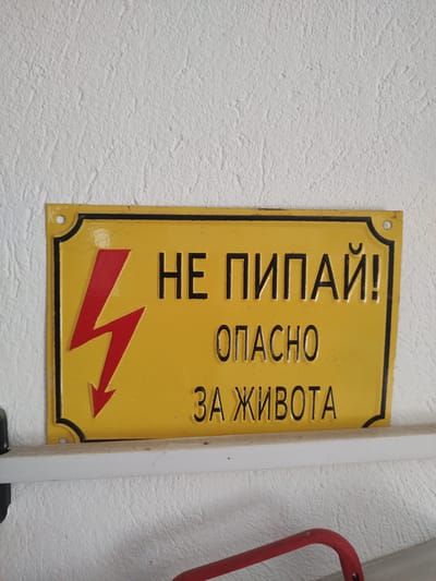 A yellow rectangular warning sign with a black border and a red lightning bolt symbol is affixed to a white, textured wall. The sign has embossed black text that reads "НЕ ПИПАЈ!" which translates to "Do Not Touch!" in Macedonian, and below that, "ОПАСНО ЗА ЖИВОТА" which translates to "Dangerous to Life". This sign is positioned on a white shelf, possibly part of an electrical enclosure, in Tervel, Bulgaria. The environment appears to be indoors, with the sign serving as a safety warning. There are no people or animals present in the image. The lighting is even, indicating a well-lit area.