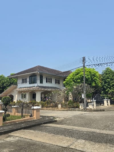 An exterior, daytime shot captures a two-story residential structure and its immediate surroundings in San Phra Net, Thailand. The house, painted a light cream or beige, features a dark tiled roof and multiple windows, some with dark frames on the upper story and others with white frames. A covered entryway with arched supports is present on the ground floor, alongside a partially visible dark-colored vehicle under a covered carport to the right. The property is fronted by a low metal fence with light-colored concrete posts, behind which a landscaped garden with various green trees and bushes is visible.
In the foreground, a gray asphalt or concrete street exhibits cracks and wear. A concrete curb separates the street from a raised sidewalk or verge. To the right, a dark utility pole supports multiple black power lines that stretch horizontally across the upper right portion of the frame. The sky above is clear and bright blue, devoid of clouds. No individuals are present, and no actions are depicted.