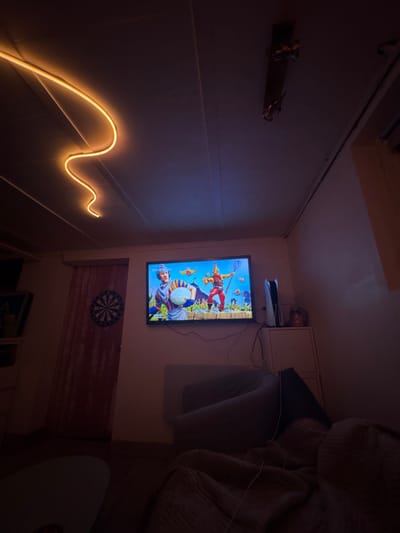 A television displays a video game scene. Above the television is a PlayStation 5. To the left of the television, an S-shaped LED strip illuminates the wall. Beneath the television is a dartboard. The right side of the view is dominated by a couch. An end table with a round, light-colored surface sits in front of the couch.