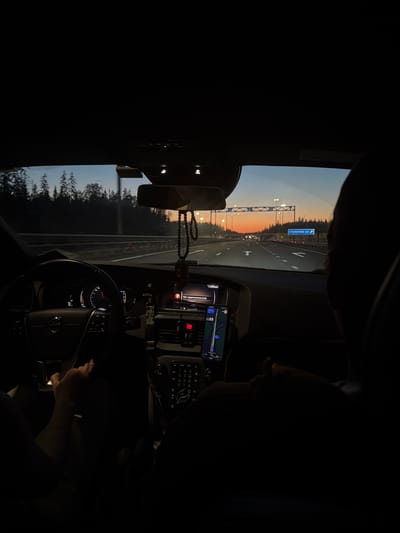The image shows the interior of a car at dusk, with a view of a highway and a sunset in the distance. The car appears to be on a highway heading towards Kissolovo, Russia, as indicated by a road sign visible in the image. Inside the car, a person's hands are visible on the steering wheel, suggesting they are driving. A phone with a navigation app is mounted on the dashboard. The overall mood is serene, with the warm colors of the sunset contrasting with the dark interior of the car.