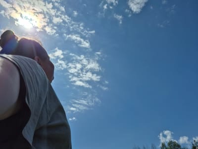 The photo is a low-angle shot looking up into a blue sky with scattered white clouds. In the lower right portion of the frame, a person's head and upper body are visible, partially obscuring the sky. The sun is behind the person's head, creating a halo effect and a rainbow-colored lens flare. The person is wearing a light gray garment. Some trees are visible in the upper left. There is no indication of the specific location being in Дойная, Russia.