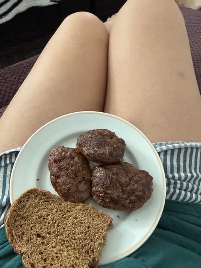 In Nogir, Russia, a person's legs are visible as they recline on a dark blanket. In front of them, on a white plate, are three browned hamburger patties and a slice of brown bread. The hamburger patties are round and slightly irregular in shape, indicating they are likely homemade. The bread appears to be whole wheat, with a visible crust and open crumb structure. The person is wearing striped pajama bottoms. The scene suggests a casual, indoor setting.