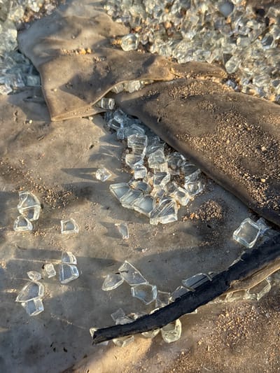The image depicts a close-up, overhead view of shattered glass pieces scattered across a rough, concrete-like surface. Two dark, flexible materials, possibly rubber or plastic sheeting, are partially visible, one draped over the glass and the other lying to the side. The scene is illuminated by direct sunlight, casting shadows across the ground and highlighting the crystalline texture of the broken glass. Small pebbles and sand are mixed in with the glass fragments, suggesting an outdoor or construction site environment. The location is identified as Quy Nhon Ward, Vietnam. No people or specific actions are depicted.