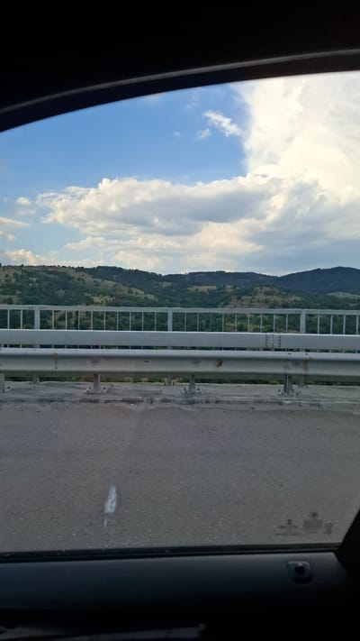 The image shows a view from inside a vehicle, looking out through a window. The foreground consists of the car's interior, including a black dashboard and part of a window frame. The middle ground features a road surface and a metal guardrail. Beyond the guardrail, a green, hilly landscape with scattered trees and some buildings is visible. The background is dominated by a bright blue sky with scattered white clouds. The scene appears to be in a rural or semi-rural area. There is no visible indication of the location being Gorno Varshilo, Bulgaria within the image itself.