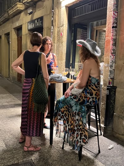 Three women are gathered outdoors in front of a building that appears to be a bar or restaurant, possibly on a street in San Sebastián, Spain. One woman, wearing a black sleeveless top and a long, colorful patterned skirt with a pink, yellow, and black geometric design, stands with her left hand on her hip. She has short, dark hair and is wearing gold hoop earrings. A green macrame bag with fringe hangs from her shoulder. Another woman, with curly brown hair, stands facing away from the camera, looking towards the first woman and the table between them. She is wearing a dark top with a pattern of small white and blue dots. The third woman is seated on a black stool, facing the other two women. She has long, curly brown hair and is wearing a grey, fuzzy hat with a red brim. Her dress features a bold animal print and a tropical floral pattern in blues, greens, oranges, and blacks. She is holding a small white crocheted bag in her lap. A small, round table is positioned between the women, with two glasses of drinks and a bottle of water on it. The background shows a building with stone walls and a dark-framed entryway. The ground is paved with textured tiles. A black trash receptacle with stickers is visible to the right of the seated woman. The lighting suggests it is evening or night.