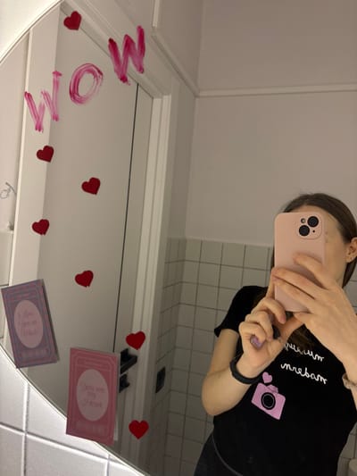 The image displays a mirror selfie taken indoors, likely within a bathroom setting. A female individual is visible in the mirror's reflection, with her face obscured by a light pink smartphone held in both hands. She wears a black short-sleeved t-shirt adorned with a pink camera graphic and partially visible white text. A black wristband or watch is present on her left wrist, and her right hand holds a key with a light purple fob. The mirror is decorated with the word "WOW" written in a thick, pink substance, consistent with lipstick, across its upper left surface. Multiple red, heart-shaped cutouts are affixed to the mirror, scattered around the text and in the lower sections. Two rectangular decorative cards are also attached to the lower left portion of the mirror: one states "I Love You so Much," and the other reads "You are my Heart." The background reflected in the mirror reveals a tiled wall with white square tiles and grey grout lines, along with a white door frame. The specific location within the city of Novosergievka, Russia, cannot be determined from the visual information provided in the image.