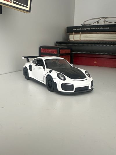 A white Porsche GT3 model car sits on a gray surface, next to a stack of books and eyeglasses. The books are of various colors and sizes. The car is slightly angled towards the right, showing its front and side details. The eyeglasses are perched on the top of the books. A small portion of a framed object is visible on the lower left of the frame. The background is a plain gray surface.