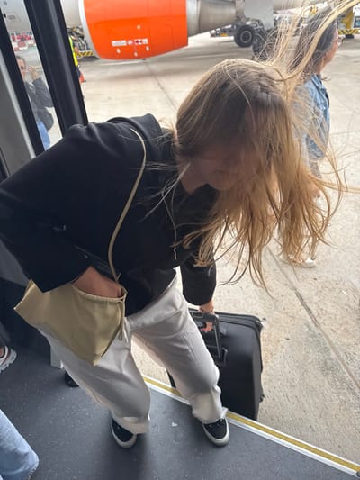 The image captures an outdoor scene at Palma de Mallorca Airport (PMI) in Son Santjoan, Spain. A fair-skinned individual with long, light brown hair, heavily affected by wind and partially obscuring their face, is shown in the process of disembarking from a vehicle, likely an airport bus or ground-level jet bridge. This individual wears a black long-sleeved top or jacket, light off-white wide-leg trousers, and black sneakers with white soles. They are actively pulling a black wheeled suitcase down dark gray steps, each marked with a yellow safety stripe. A light beige fabric shoulder bag with a drawstring closure is worn across their body, with their left hand inserted into it. In the background, a prominent orange jet engine nacelle, characteristic of an EasyJet aircraft, is visible on the concrete tarmac, along with a portion of the white fuselage and landing gear. To the right, partially visible, another fair-skinned individual with dark hair, wearing a denim jacket and light-colored trousers, stands on the tarmac, facing away from the camera, their hair also appearing wind-blown. A blurred reflection of a third person is faintly discernible in the vehicle's doorframe on the far left.