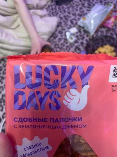 The image shows a pink package of "LUCKY DAYS" cookies with purple striped lettering. The cookies are described as "сдобные палочки с земляничным джемом" (fluffy sticks with strawberry jam) and promise "сладкое удовольствие" (sweet pleasure). A white thumbs-up graphic is also visible. The packaging has a red, serrated edge. In the background, a person's arm and hand are visible, along with leopard-print fabric and other snack packaging. The image does not contain any information about the city of Zmeinyi, Russia.