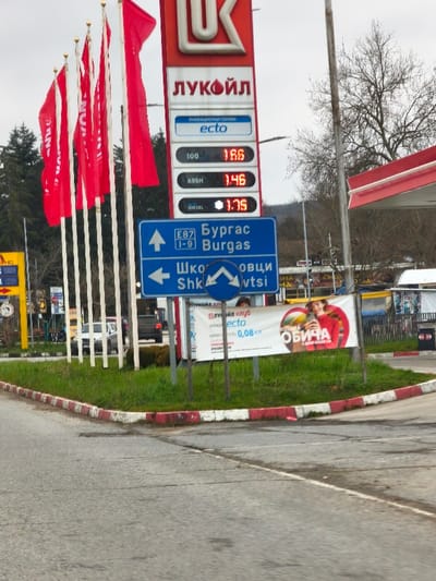 The image displays a roadside scene at a Lukoil gas station in Staro Oriahovo, Bulgaria, under an overcast sky. A tall, red-and-white Lukoil sign is centrally featured, displaying fuel prices: "100" at 1.86, "A95H" at 1.46, and "DIESEL" at 1.75, presumably in Bulgarian Leva. To the left of the sign, multiple red flags on white poles are arranged in a row. In the foreground, a grassy median with a red-and-white painted curb separates the road from the gas station entrance. A blue rectangular road sign directs traffic straight for "E87 I-9 Бургас Burgas" and left for "Шкорпиловци Shkorpilovtsi" with a U-turn symbol. Below this, a circular blue sign mandates a left U-turn. Two white advertising banners are positioned beneath the road signs; one mentions "ЛУКОЙЛ клуб" and "ecto 0,08," while the other depicts a smiling woman holding a smartphone. In the background, a silver SUV and a dark pickup truck are visible on a road. A red-and-white gas station canopy is partially visible on the right, and various trees, including evergreens and a bare deciduous tree, frame the scene. No individuals are actively present in the depicted environment, only portrayed on an advertisement.