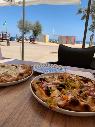 Two pizzas are on a wooden table in front of a window. The pizza in the foreground is topped with a creamy white sauce, ham, and some green herbs. The pizza in the background has a red sauce base with toppings of mushrooms, olives, red onion, yellow and green bell peppers, and capers. A blue and white striped plate sits between the two pizzas.

Through the window, a beach scene is visible. The sky is a clear blue, indicating it is daytime and sunny. There are some trees and beach umbrellas. In the distance, there are people on the beach and some buildings. The word "HOTEL" is partially visible on a white umbrella to the left of the frame. The scene suggests a casual meal at a restaurant with a view of the beach.