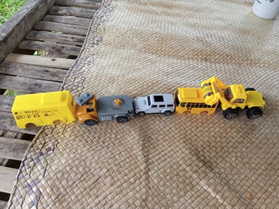 The image shows a line of toy vehicles on a woven mat, which is partially resting on a weathered wooden bench. From left to right, the toy vehicles include a yellow school bus, an orange semi-truck cab, a gray flatbed trailer, a white SUV, another yellow school bus, and a yellow construction excavator. The background suggests an outdoor setting. The image does not contain any people or actions. The location in the city of Palohigeuh, Indonesia is not verifiable from this image alone.